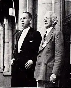 Vue de près de deux hommes en costume, l'un des deux est âgé, devant un bâtiment.