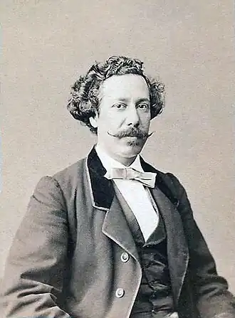 Photographie en noir et blanc. Portrait en buste d'un homme élégant avec les cheveux bouclés et une moustache dont les pointes remontent. Il porte un nœud papillon et un costume trois-pièces.