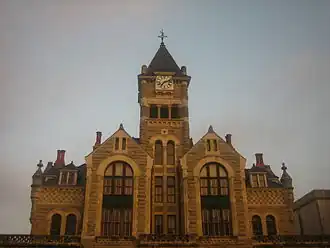 Vue d'un bâtiment à la façade de pierres avec, au centre, une tour surmontée d'un clocher avec horloge.