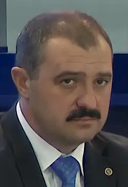 Plan serré d'un homme moustachu habillé en costume et cravate bleu.