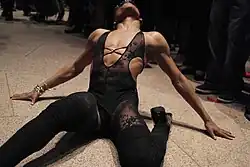 Photographie d'un danseur noir en train de voguer. Mains sur le sol, mollet replié sous sa cuisse, tête renversée, il porte une tenue noir moulante en dentelle.