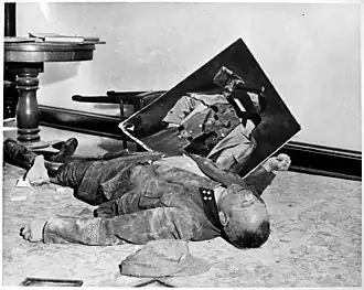 Photo noir et blanc prise le 19 avril 1945, à l'intérieur d'un bureau de l'hôtel de ville de Leipzig. Sur le sol, un homme mort, en uniforme, est allongé de tout son long, sa tête vers la droite au premier plan. Un mur blanc forme l'arrière-plan de la photo. Au second plan, entre une chaise renversée le long du mur et le flanc droit du cadavre, tient en équilibre un portrait d'Adolf Hitler dont le visage a été lacéré.