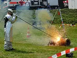 Un homme vêtu de manteaux protecteurs tient une longue tige d'où jaillit une boule de feu contre un bloc de métal.