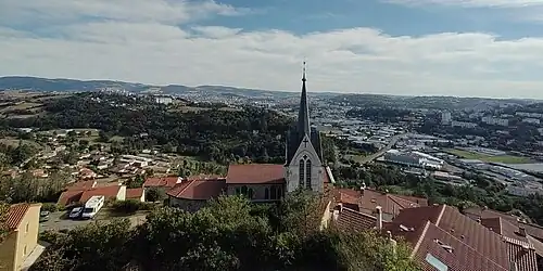 On peut voir le clocher de l'église du village parmi de nombreux toits et, à l'arrière plan, le nord de la ville de Saint-Étienne dont le coeur est dissimulée par la verdoyante colline de Montreynaud.
