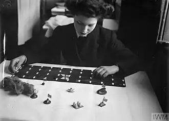 Une Wren préparant du matériel pour un wargame. Les pièces en bois représentent des escorteurs. La planche noire avec des formes blanches simule le convoi. L'écheveau de laine est destiné à simuler les écrans de fumée.