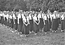 Photographie en noir et blanc d'un groupe de jeunes filles debout en uniforme et chemise blanche. Disposées géométriquement, elles se tiennent au repos.
