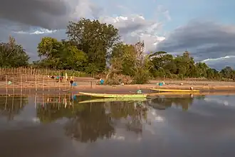 Réflexion (optique) sur l'eau d'une berge avec des arbres et deux pirogues au coucher du soleil sur l'île de Don Puay à Si Phan Don.