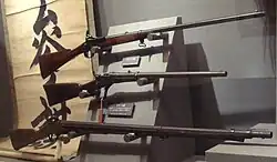 Photographie d'un présentoir de musée sur lequel sont placés trois fusils de forme et de taille différentes, la crosse à gauche et le canon à droite