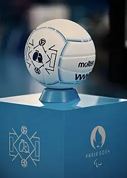 Gros plan sur un ballon blanc avec des liserés bleus, avec le nom de la marque et le logo du rugby-fauteuil de Paris 2024 en noir. Le ballon est posé sur un stand de présentation avant un match, avec sur un côté le logo du rugby-fauteuil et sur un autre côté le logo de Paris 2024 et les Agitos.