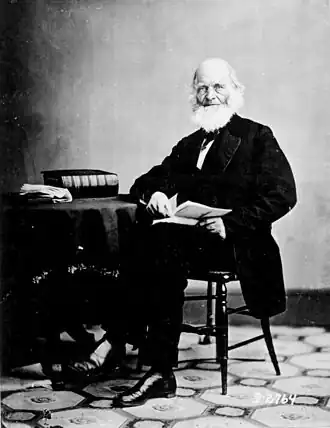 La photographie noir et blanc montre un homme âgé dans un costume noir. Il porte une barbe blanche imposante, et tient un livre ouvert, assis sur une chaise. Il est accoudé à une table sur laquelle repose un épais livre noir et une liasse de papier.