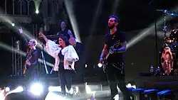 Photo en couleur d'un groupe de musique sur une scène, une chanteuse brune habillée de blanc lève un bras