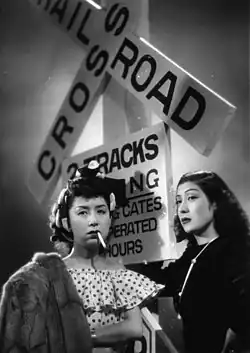 Deux femmes se tiennent aux côtés d'un poteau portant des pancartes, sur l'une d'elles on peut lire « Railroad Crossing ». La première, une prostituée (Kinuyo Tanaka) se tient les bras croisés, un manteau de fourrure posé sur une épaule et une cigarette allumée pendue aux lèvres, la seconde (Sanae Takasugi) en robe noire a la main posée sur le poteau (image en noir et blanc).