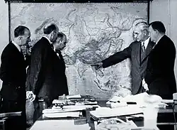 Photo en noir et blanc montrant cinq hommes en costume regardant une carte murale de l'Asie. L'un des hommes pointe la région du Bangladesh à l'Est de l'Inde. Au premier plan, un bureau est recouvert de documents.