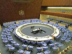 Une grande pièce feutrée avec au centre un bureau blanc simple à deux places équipé d'un ordinateur portable et quelques appareils de communications. Le bureau est entouré d'un grand bureau circulaire pouvant accueillir entre 30 et 40 personnes. Le bureau circulaire dispose de micro (une trentaine). Une quarantaine de chaises studieuses bleues sont placés autour du bureau circulaire. Le tout est entourés de deux autres cercles concentriques de chaises, identiques aux premières. Sur les côtés, des rangés de chaises permettent d’accueillir plusieurs dizaines de personnes supplémentaires, certaines en hauteur dans des loges. La pièce est dominée par un immense mur en bois sur lequel on voit très clairement le logo de l'OMS.