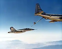 Un avion de chasse vole en arrière d'un avion ravitailleur dont le perche de ravitaillement est déployée ; le contact entre les deux avion n'est pas encore établi.