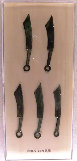 Photographe montrant cinq objets en forme de couteau derrière une vitrine. De formes similaires, les objets sont divisés en deux rangées de 2 et 3 couteaux.