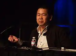 Homme brun d'origine asiatique habillé d'un costume noire et d'une chemise claire.