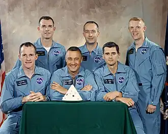 Photographie en couleur de six hommes en combinaison de vol posant avec une maquette de capsule Apollo.