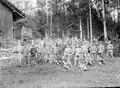 Photographie noir/blanc d’hommes, en uniforme militaire pour la plupart, posant en groupe près d’une grange en bois et devant une forêt
