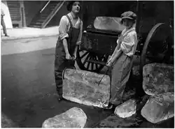 Photographie de deux jeunes femmes en salopettes tenant un bloc de glace avec des pinces. Elles se trouvent à l'arrière d'une charrette à côté de plusieurs blocs de glace déjà déchargés sur la chaussée.