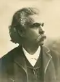 Dessin en buste d'un homme moustachu en costume sombre, aux longs cheveux bruns rejetés en arrière et au regard fiévreux.